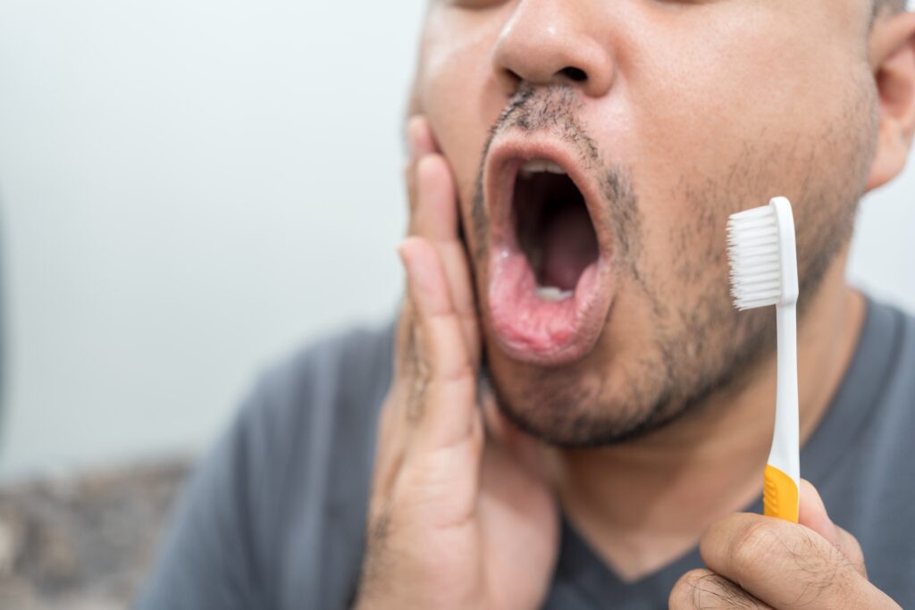 closeup of a man brushing his teeth in the mirror, holding his jaw in pain